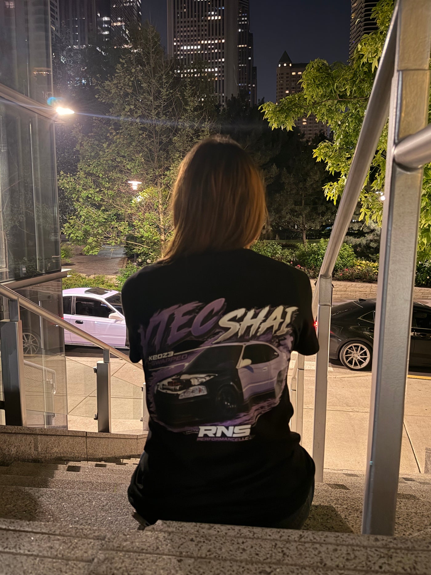 A person sitting on outdoor concrete stairs at night, viewed from behind, wearing a black graphic t-shirt with a purple and white car design and the text 'VTEC SHAI' 

